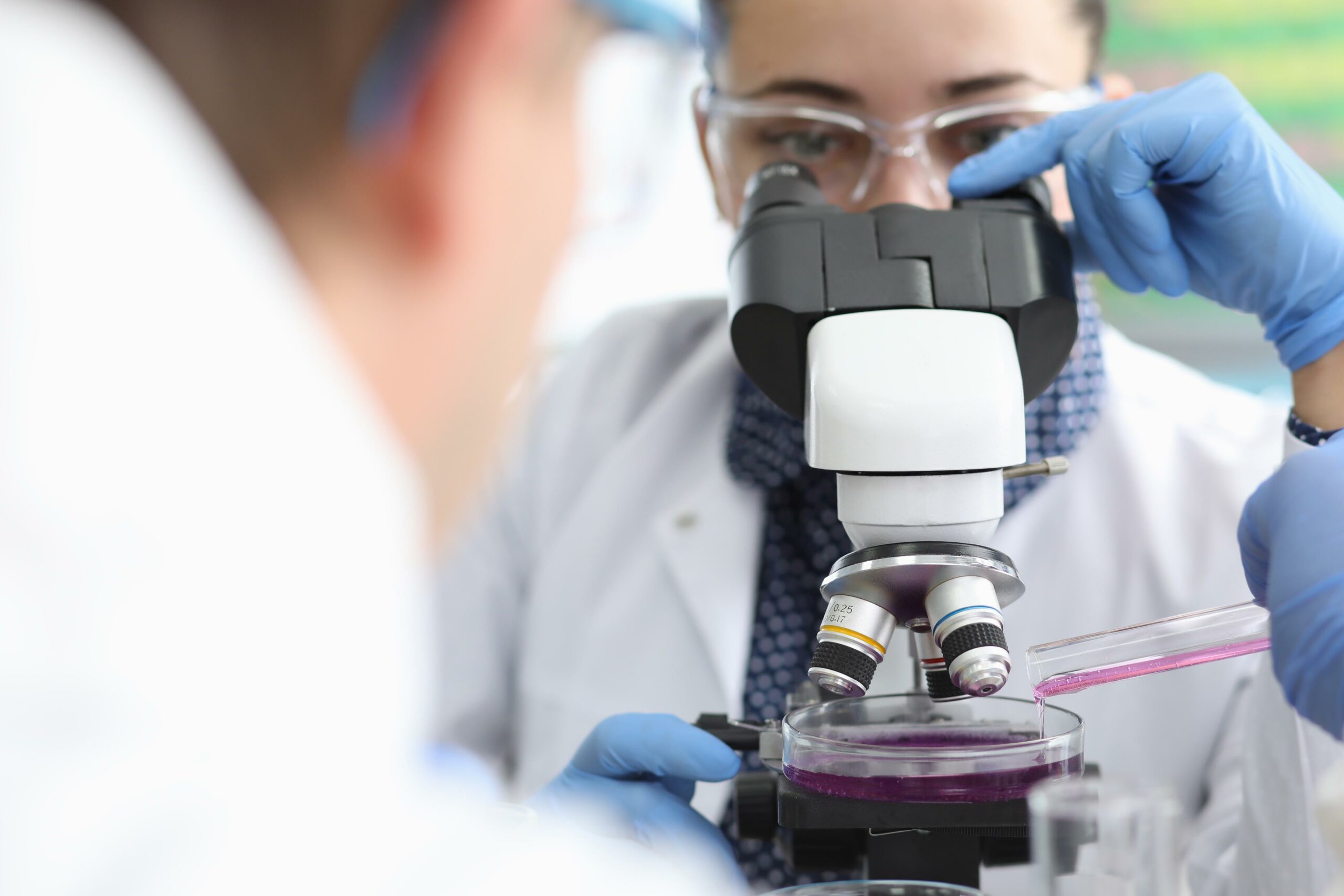 Close-up of female doctor analyzing material under microscope. Male worker adding liquid in glass round container. Laboratory tests and science concept